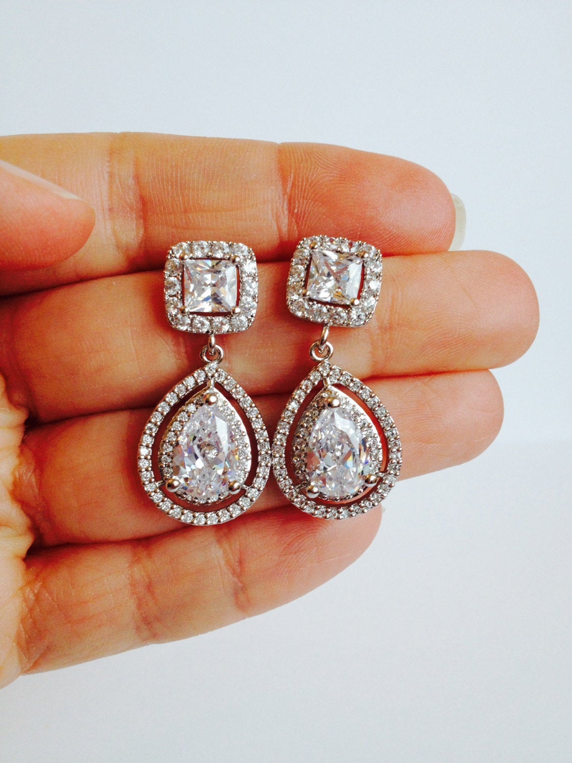 Hand holding Cubic zirconia teardrop crystal earrings in a silver colored rhodium plated brass setting.