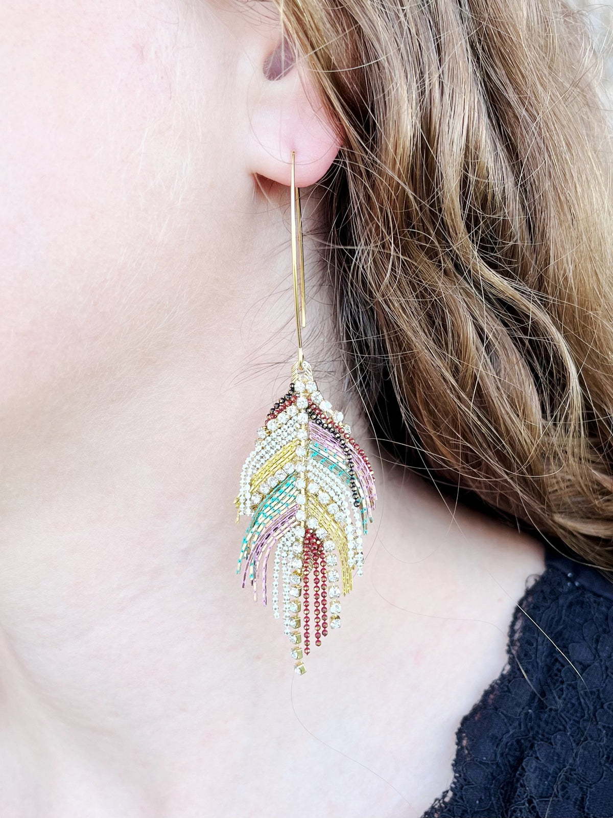 A close-up of a person's ear wearing a long, dangling feather-shaped earring made of gold-toned metal and multicolored rhinestones.