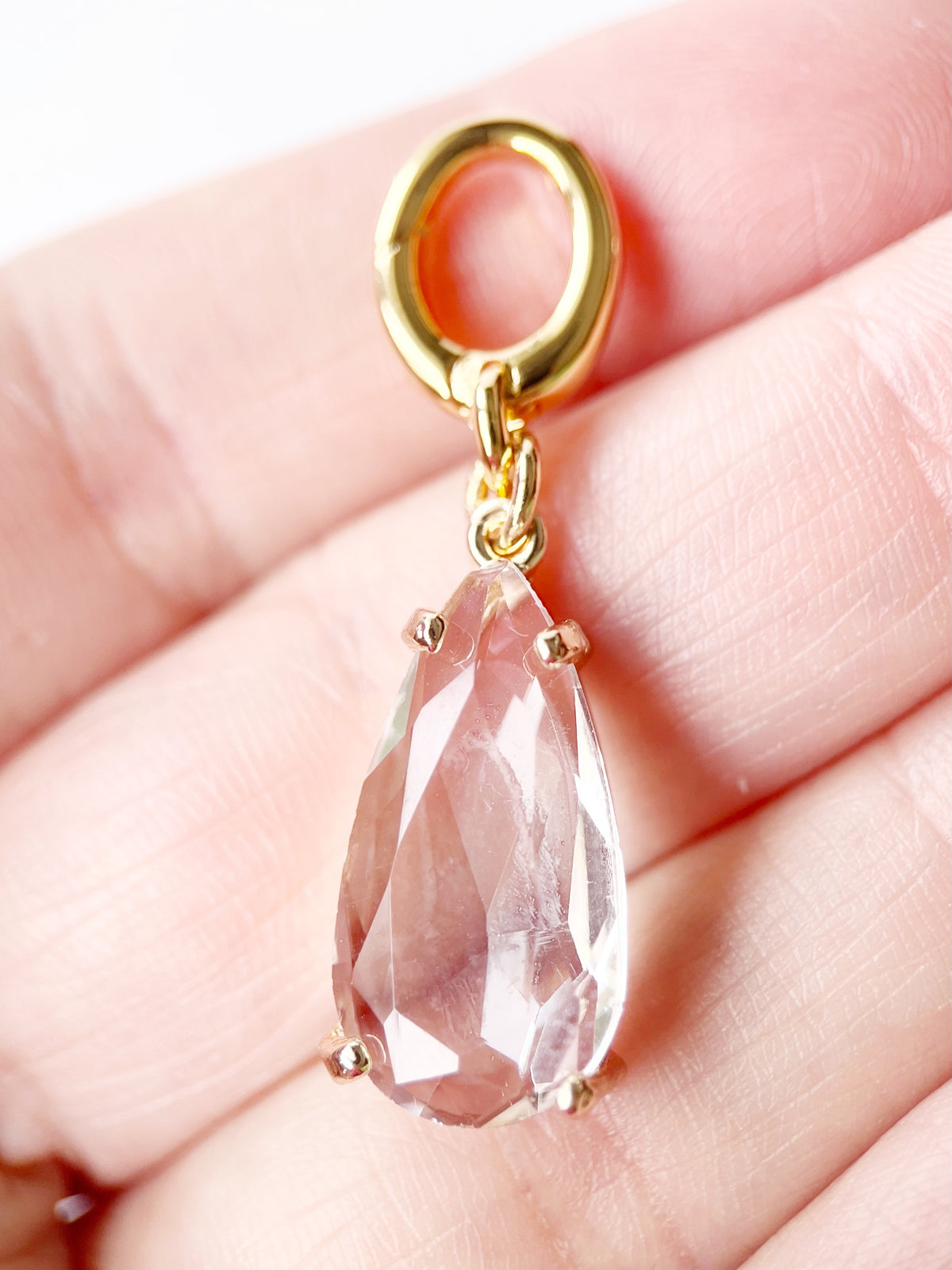 A close-up of a gold teardrop pendant with a faceted clear stone, held in a person's hand.
