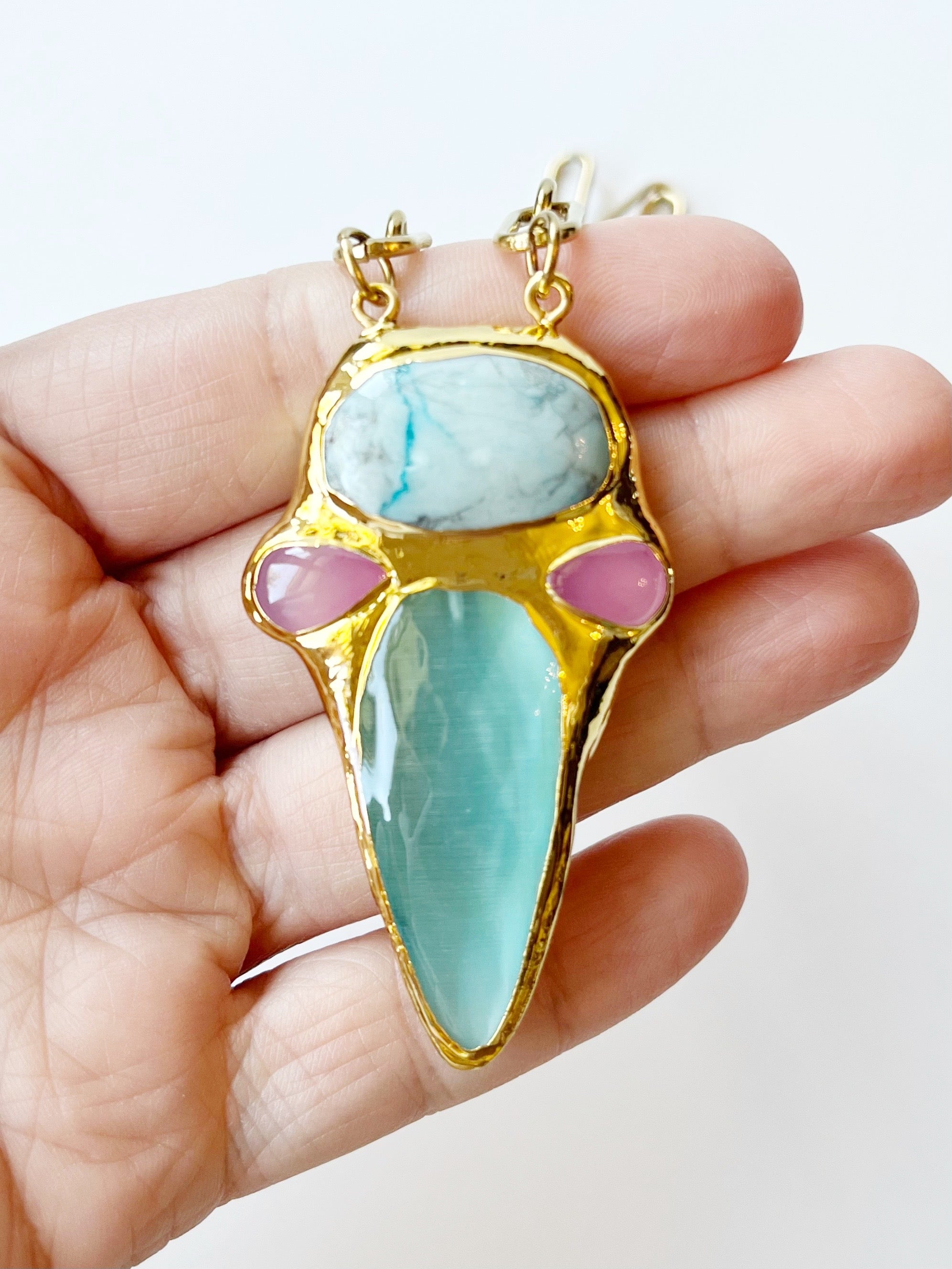 Gold pendant with turquoise and pink stones held in a hand against a white background