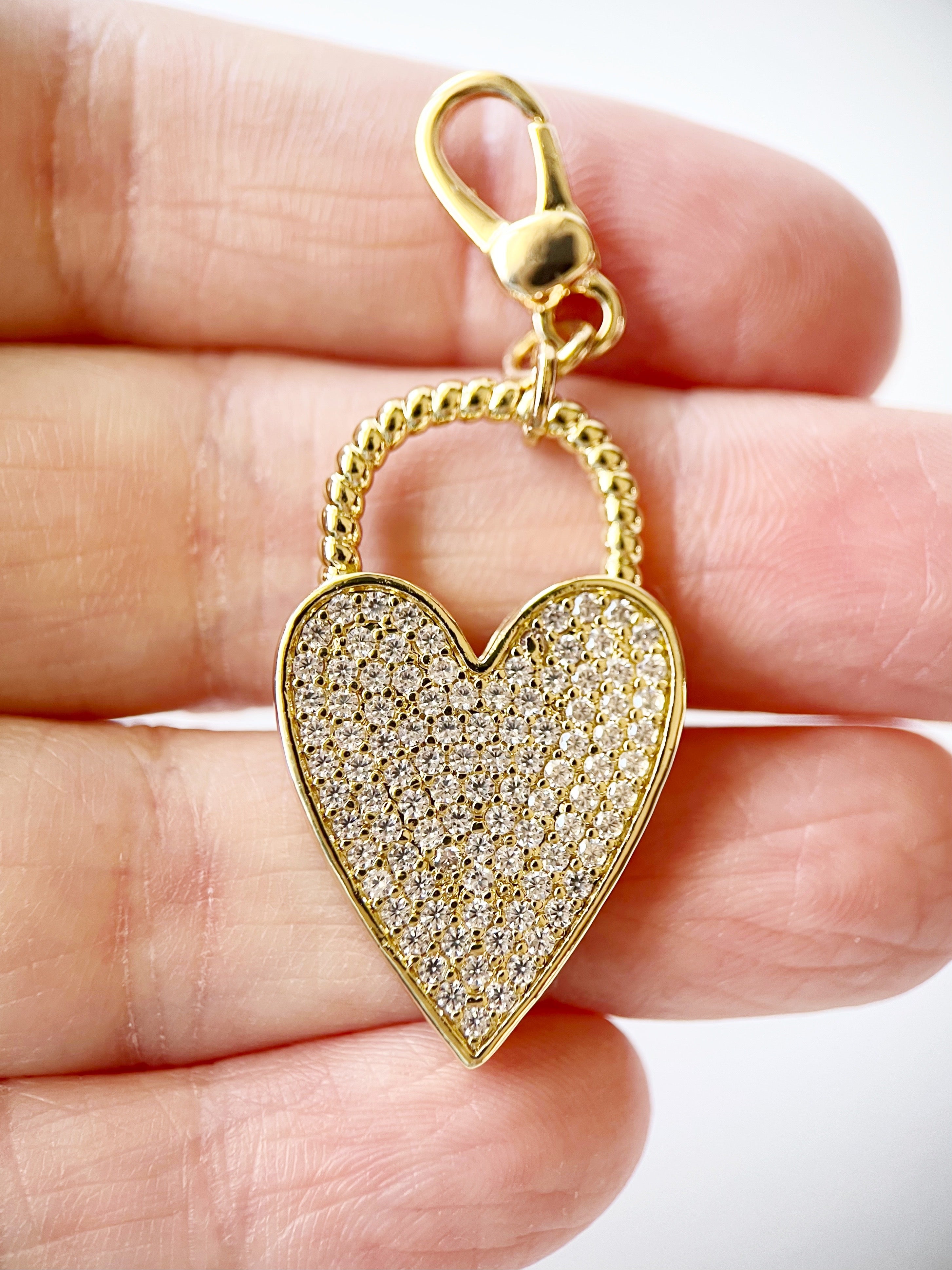 Gold heart-shapedcharm with clear stones held between fingers on a white background