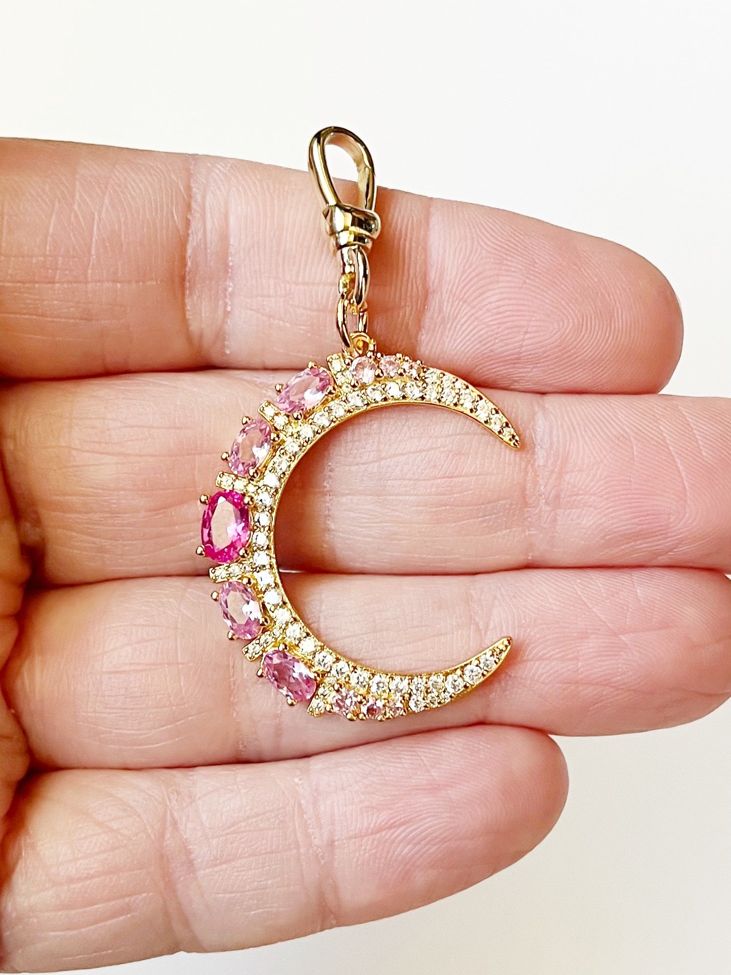 Gold crescent moon charm with pink gemstones held between fingers on a light background
