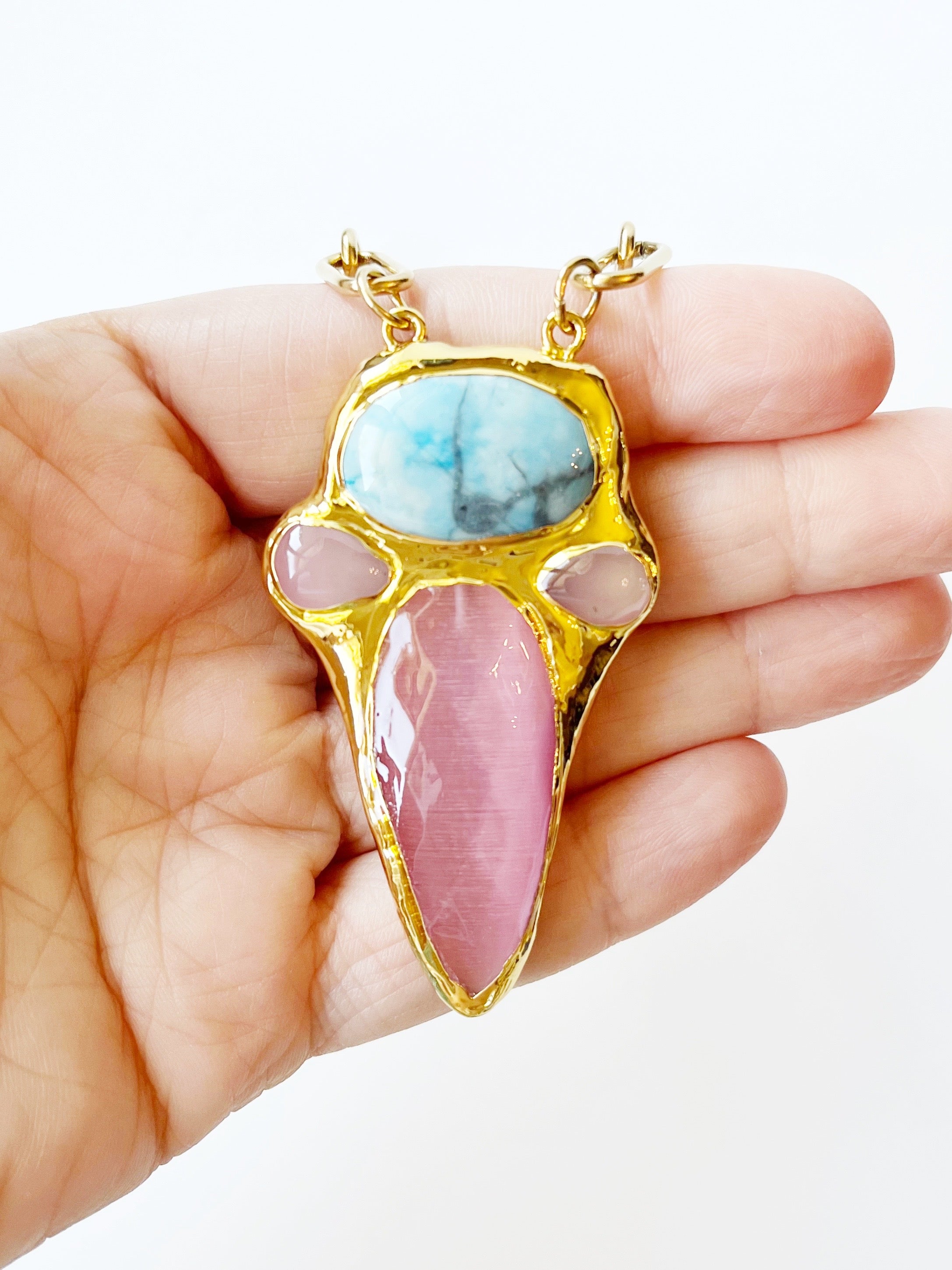 Gold pendant with pink and blue stones held in a hand against a white background