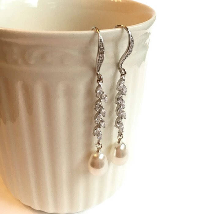 Glass pearl earrings accented by clear cubic zirconia crystals in a silver colored rhodium plated brass setting hanging on side of white mug