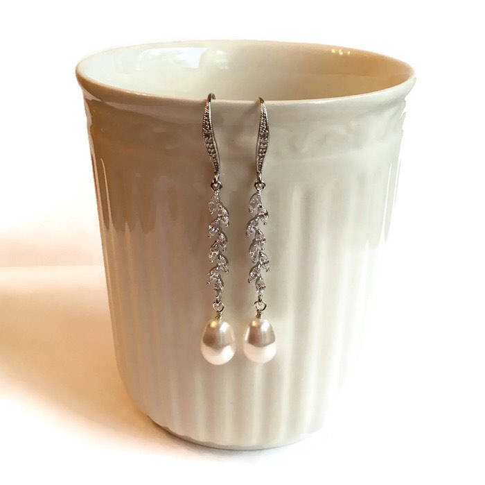 Glass pearl earrings accented by clear cubic zirconia crystals in a silver colored rhodium plated brass setting handing from white coffee mug.