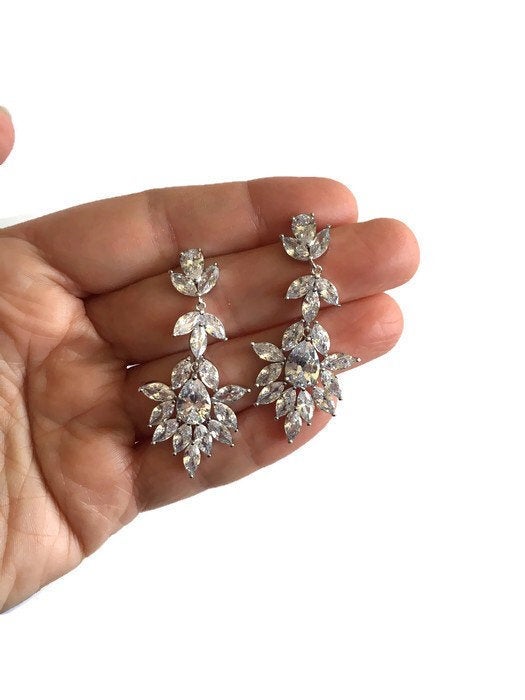 Hand holding Long Cubic zirconia crystal drop earrings in a silver colored rhodium plated brass setting.