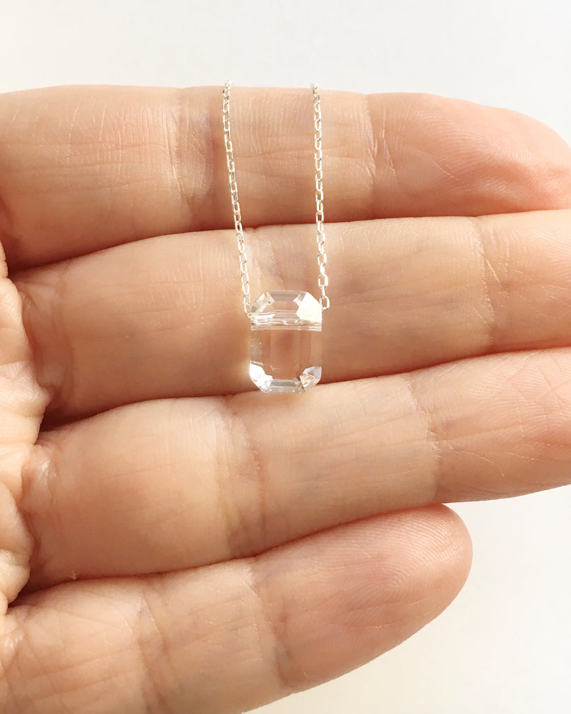 Small Clear Crystal pendant on Silver chain Necklace held across women's fingers.