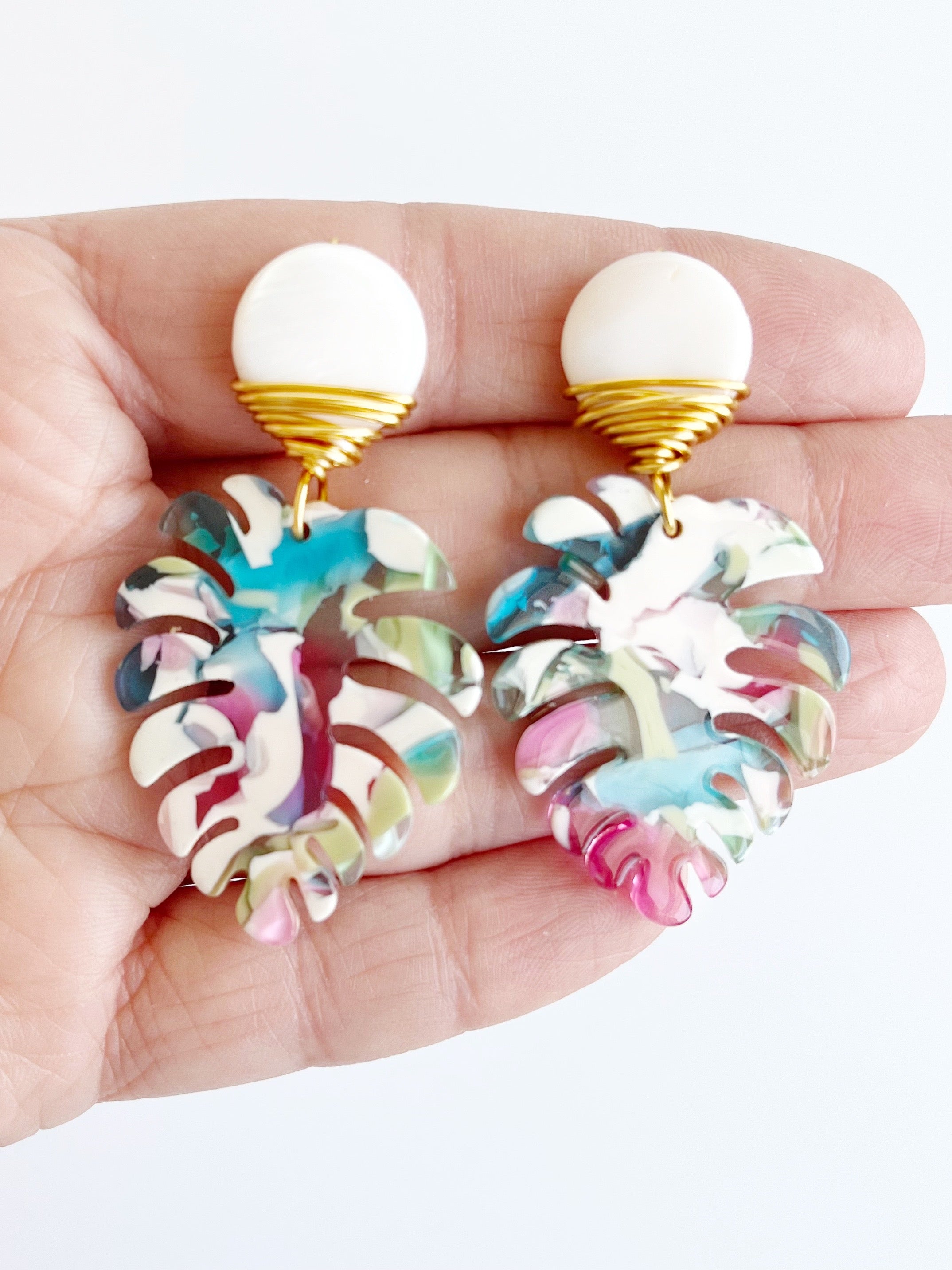 Hand holding Mini Palm Dangle Leaf Earrings in Blue, Pink, and White wrapped with gold plated wire earrings