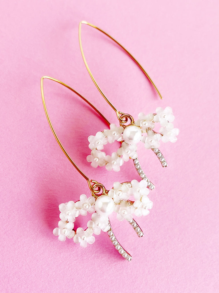 Long gold dangle earrings with pearl and flower bow pendant on a pink background.