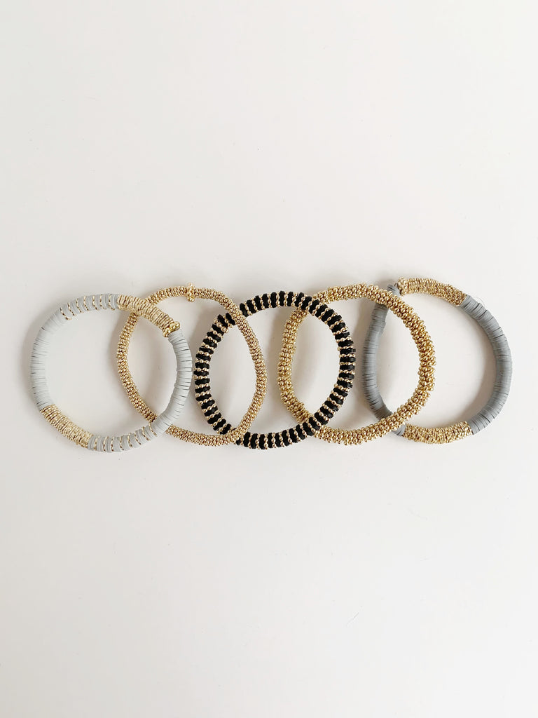 Bundle of bracelets laying over lapping they are -light gray confetti bracelet -small gold confetti bracelet -medium gold confetti bracelet -dark gray confetti bracelet -black and gold (the rosella) confetti bracelet