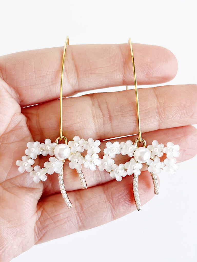 Long gold dangle earrings with pearl and flower bow pendant handing on women's hand across her fingers.