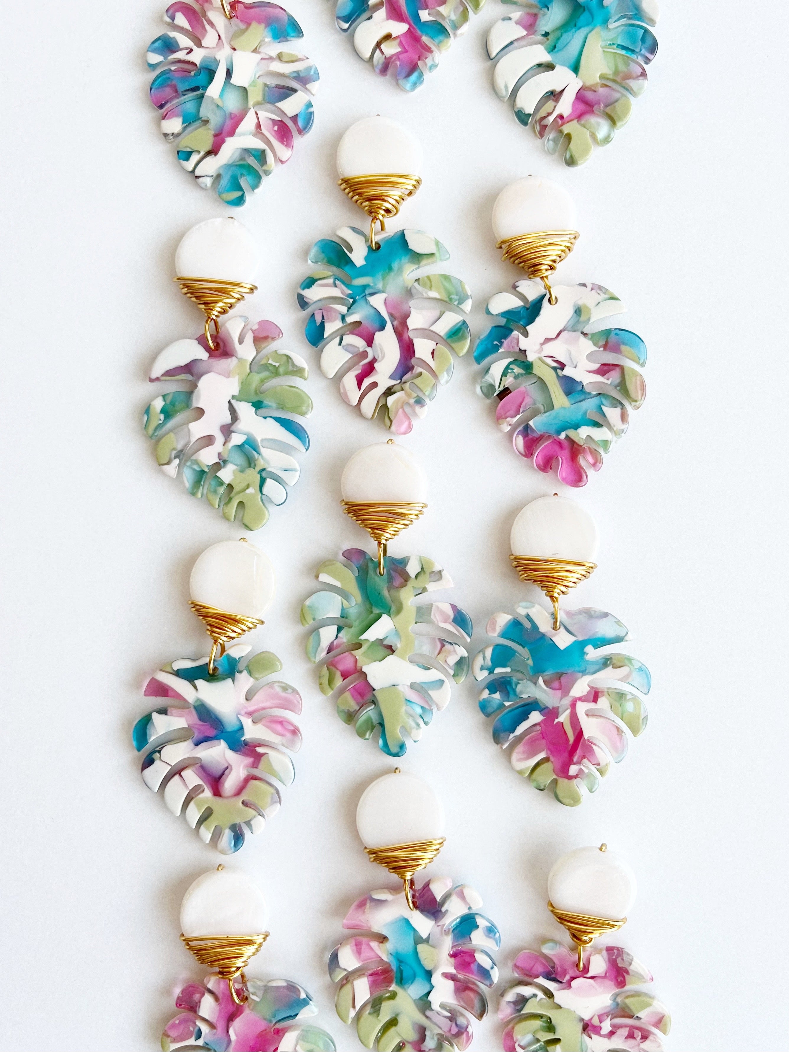 Lots of Mini Palm Dangle Leaf Earrings in Blue, Pink, and White wrapped with gold plated wire earrings on table.