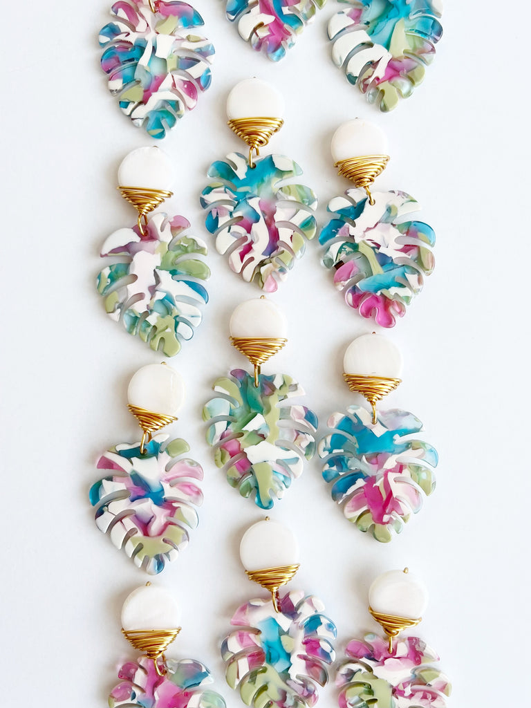 Lots of Mini Palm Dangle Leaf Earrings in Blue, Pink, and White wrapped with gold plated wire earrings on table.