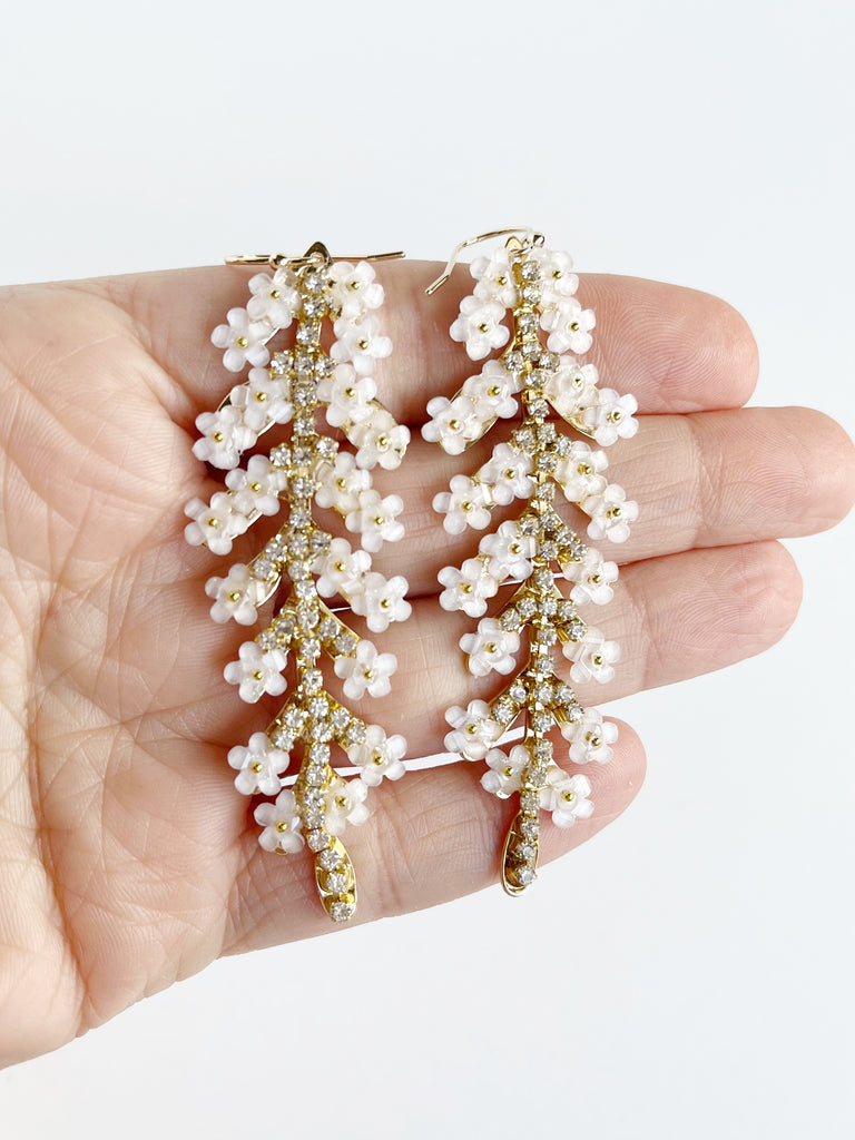 leaf shaped gold statement earrings with crystals and white flowers displayed on hand